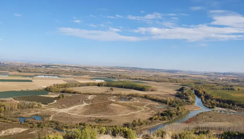 Paisaje rural con un río serpenteante cerca de Villafranca del Bierzo. Campos cultivados y árboles de otoño bajo un cielo azul con nubes.