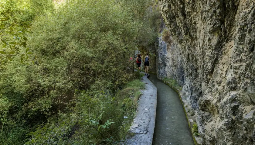 Senderistas caminan por la ruta de los bolos, un sendero estrecho junto a un canal de agua y un acantilado rocoso.
