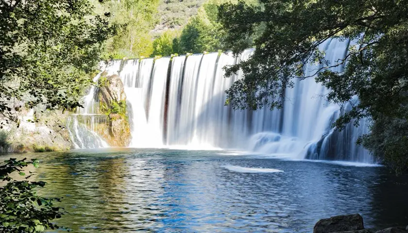 El Salto del Pelgo, una cascada imponente con agua sedosa cayendo sobre rocas, rodeada de vegetación exuberante y un estanque azul profundo.