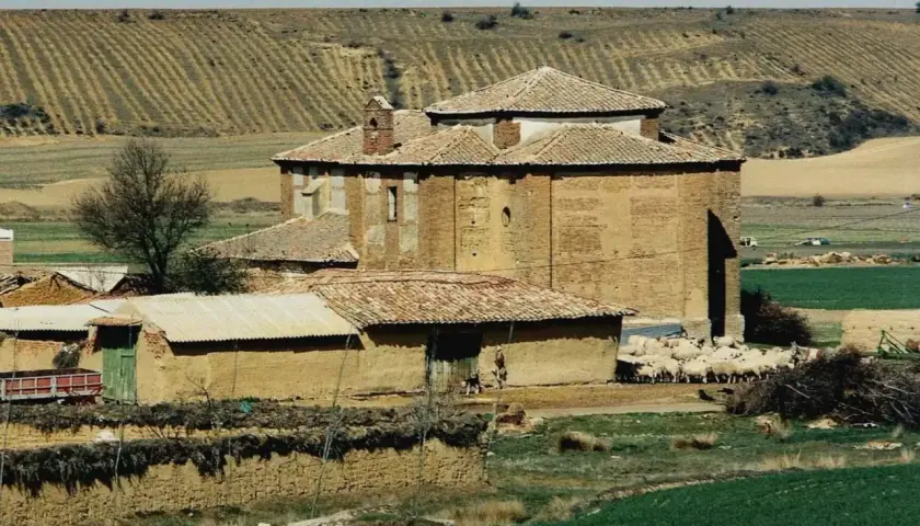 Edificio de ladrillo con tejado a dos aguas, rebaño de ovejas pastando cerca. Paisaje rural con campos de cultivo en terrazas, típico de calzadilla de la cueza.