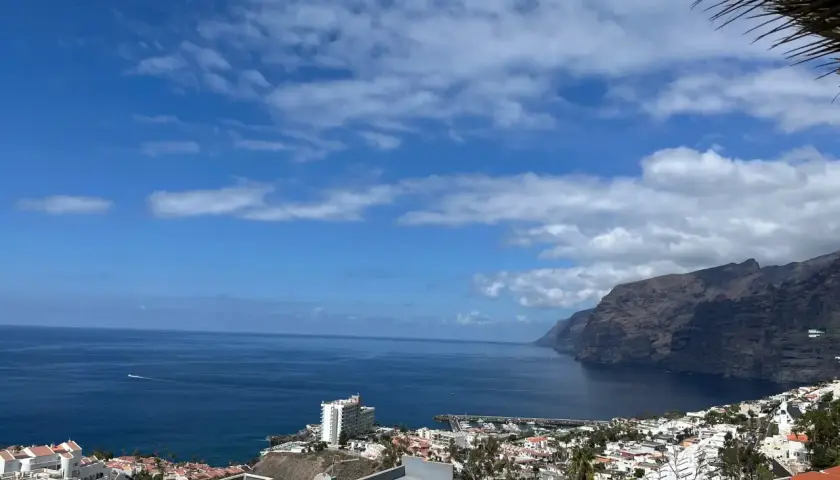 Vista panorámica de acantilados imponentes y el mar azul, con un pueblo costero y un puerto deportivo. Un lugar que evoca las mejores playas de Tenerife.