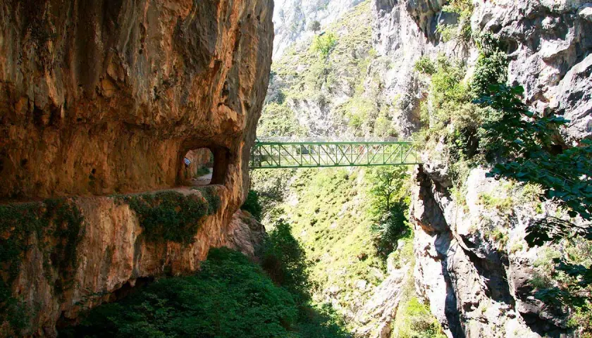 Un puente verde cruza un desfiladero rocoso, parte de la ruta del Cares. El tiempo parece perfecto para esta aventura.