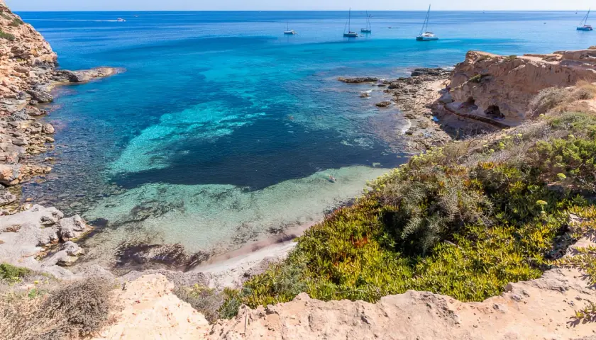Aguas turquesas y arena blanca en una cala rocosa, un paraíso entre las mejores playas de Formentera. Yates navegan en el horizonte.