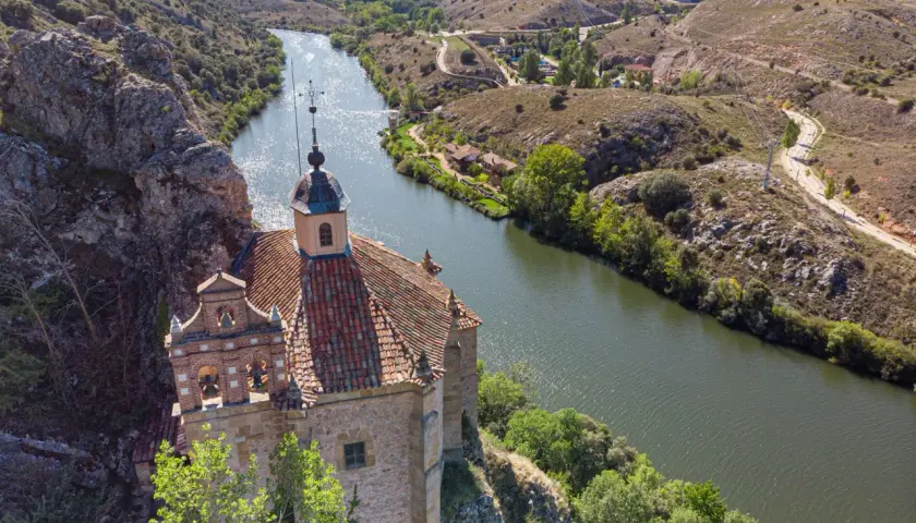 Ermita de San Saturio, Soria, se alza sobre un acantilado junto al río Duero, rodeada de vegetación y colinas.