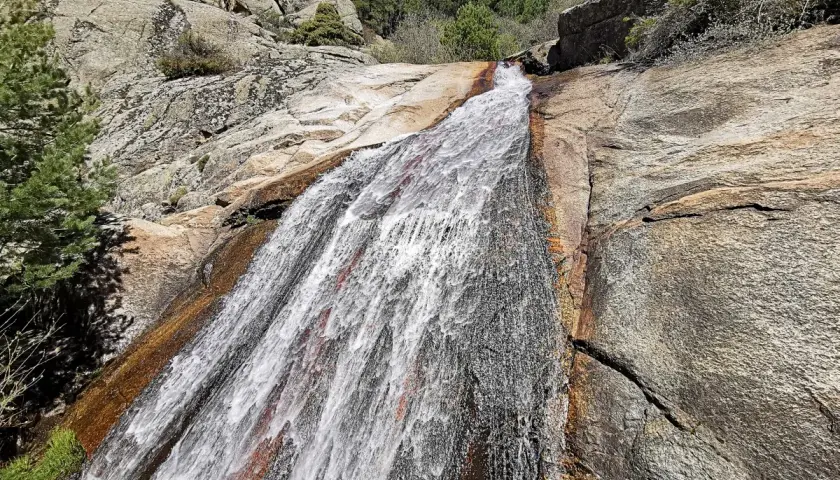 El agua desciende por la roca en la cascada del Hornillo, rodeada de vegetación y rocas graníticas bajo un cielo azul.