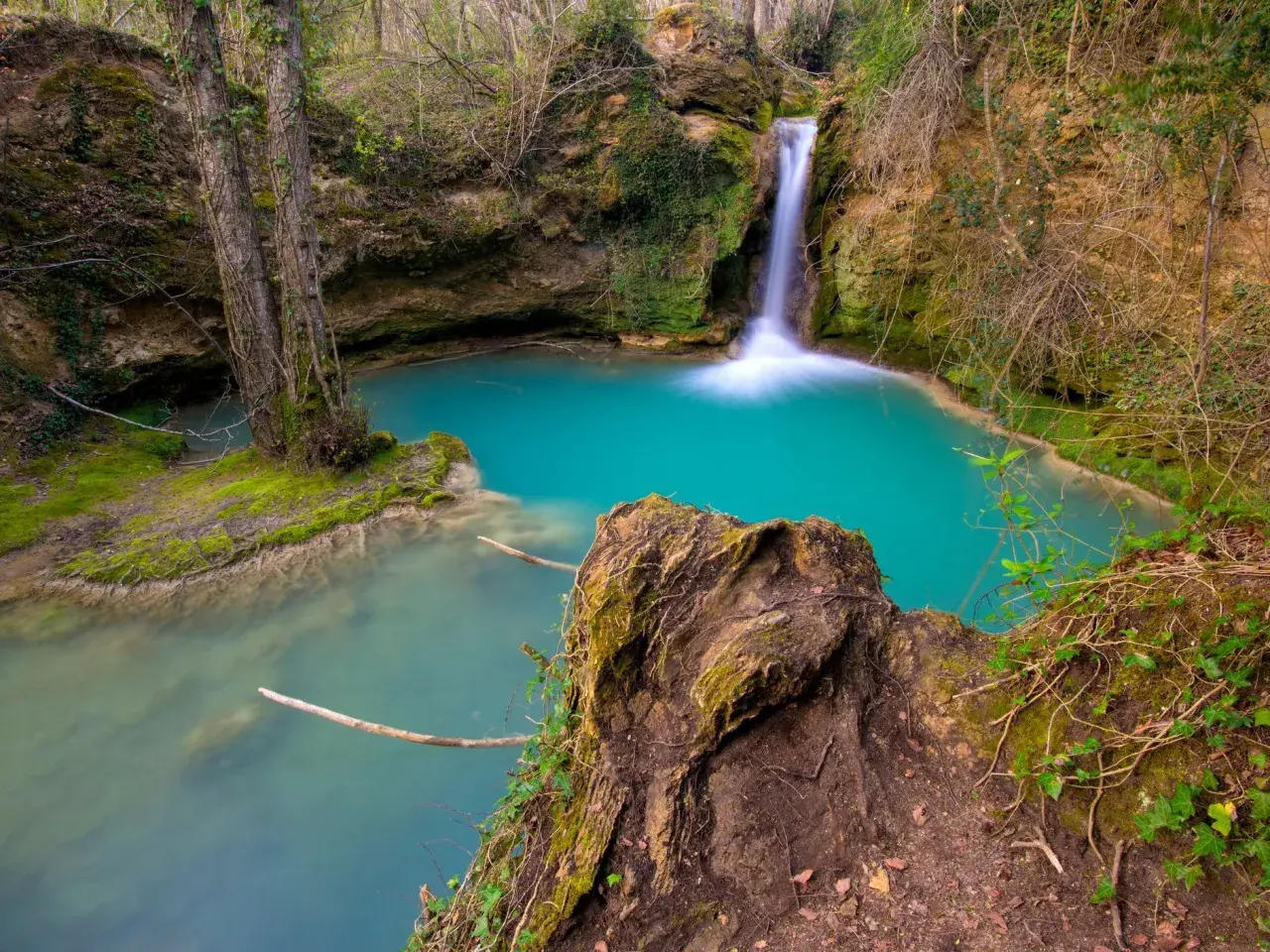 Cascada de agua turquesa en la ruta del agua Berganzo, rodeada de vegetación y rocas cubiertas de musgo.