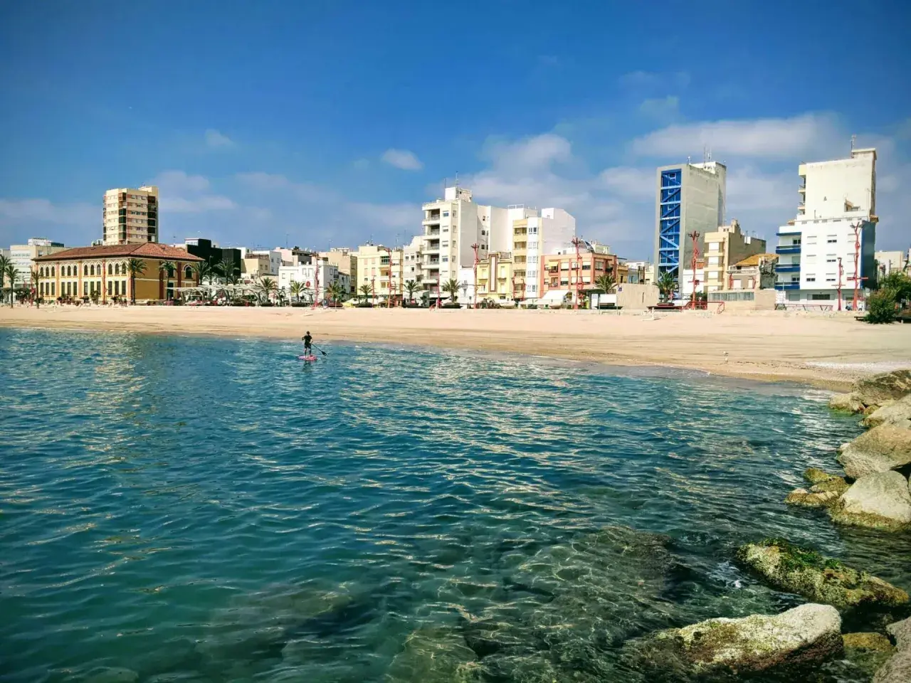 Un día soleado en la costa, con un paddleboarder solitario en el mar turquesa y edificios modernos de fondo. ¡Un oasis de playas cerca de Zaragoza!