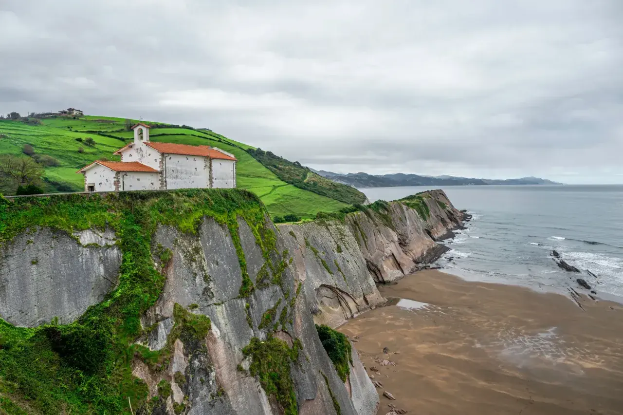 Iglesia blanca con tejado naranja sobre acantilado verde, junto al mar Cantábrico. Un lugar que evoca la esencia de "8 apellidos vascos".