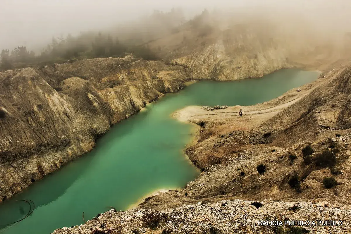 Laguna turquesa en el monte Neme Carballo, rodeada de tierra y rocas, con niebla al fondo.