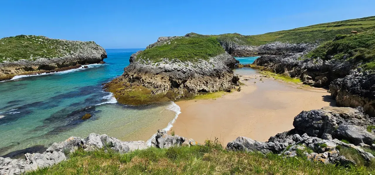 Una cala escondida entre acantilados verdes, con aguas turquesas y arena dorada. Un rincón idílico entre las mejores playas de Asturias.
