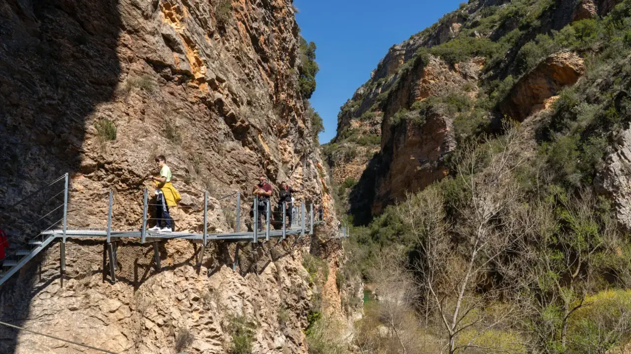 Gente camina por las pasarelas de Alquézar, un sendero suspendido entre imponentes paredes rocosas y vegetación.