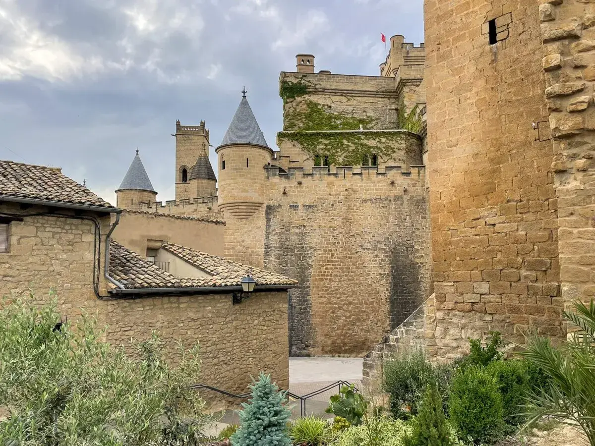 El **castillo de Olite**, con sus torres cónicas y muros de piedra, se alza majestuoso bajo un cielo nublado. En primer plano, tejados de tejas y vegetación verde añaden un toque de vida.