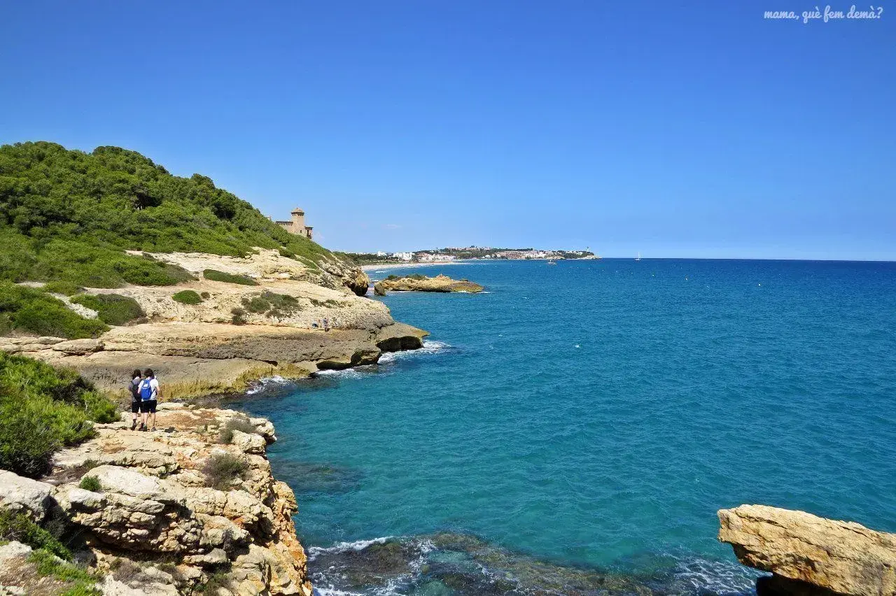 Dos personas caminan por el camí de ronda de Tarragona, disfrutando de las vistas al mar azul y la costa rocosa.