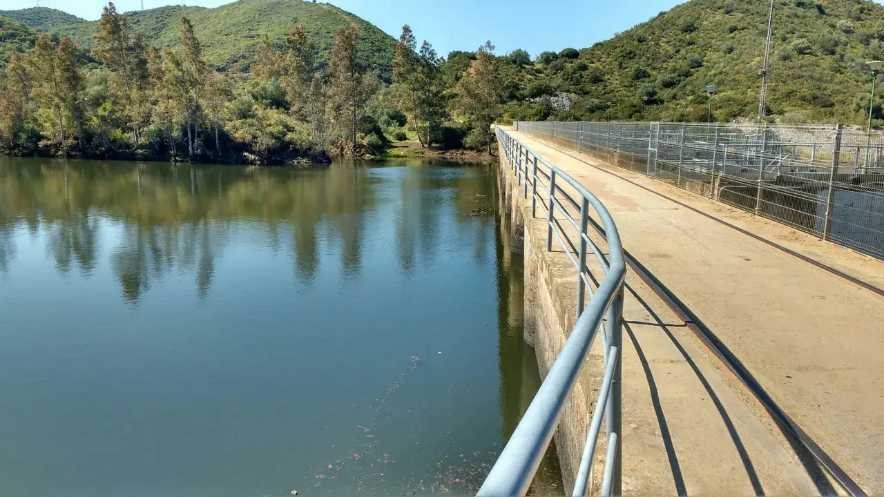 Un puente de hormigón con barandilla recorre un embalse, ideal para una ruta senderismo Sevilla. El agua refleja árboles y colinas verdes.