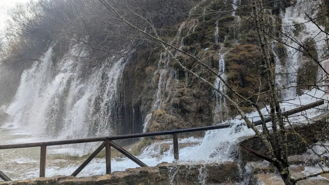 Cascada en la ruta nacimiento río Cuervo. Agua cayendo sobre rocas cubiertas de musgo y vegetación. Barandilla de madera en primer plano.