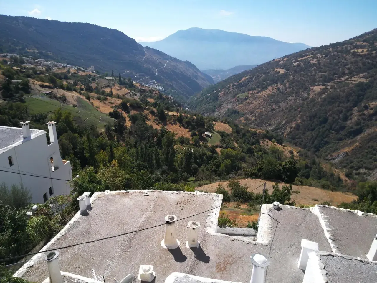 Paisaje montañoso con casas blancas y tejados planos. El camino a Trevélez se abre paso entre verdes colinas y valles profundos bajo un cielo azul.