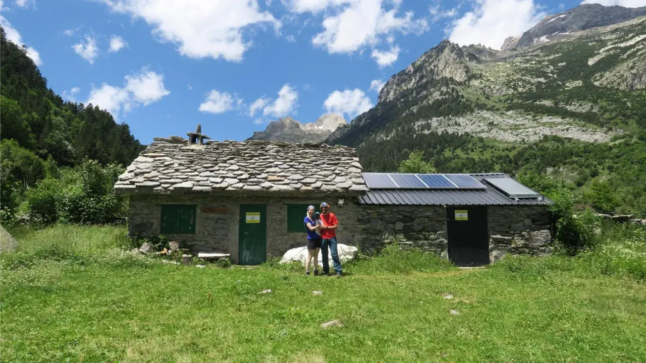 Pareja posa frente a una rústica cabaña de turmo con paneles solares, rodeada de exuberante vegetación y majestuosas montañas.