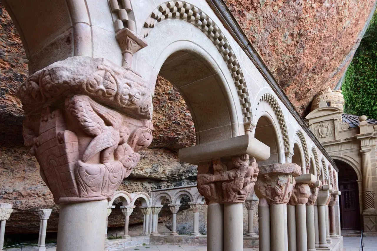 Detalles arquitectónicos y escultóricos en piedra de San Juan de la Peña, con arcos y capiteles tallados bajo una roca.