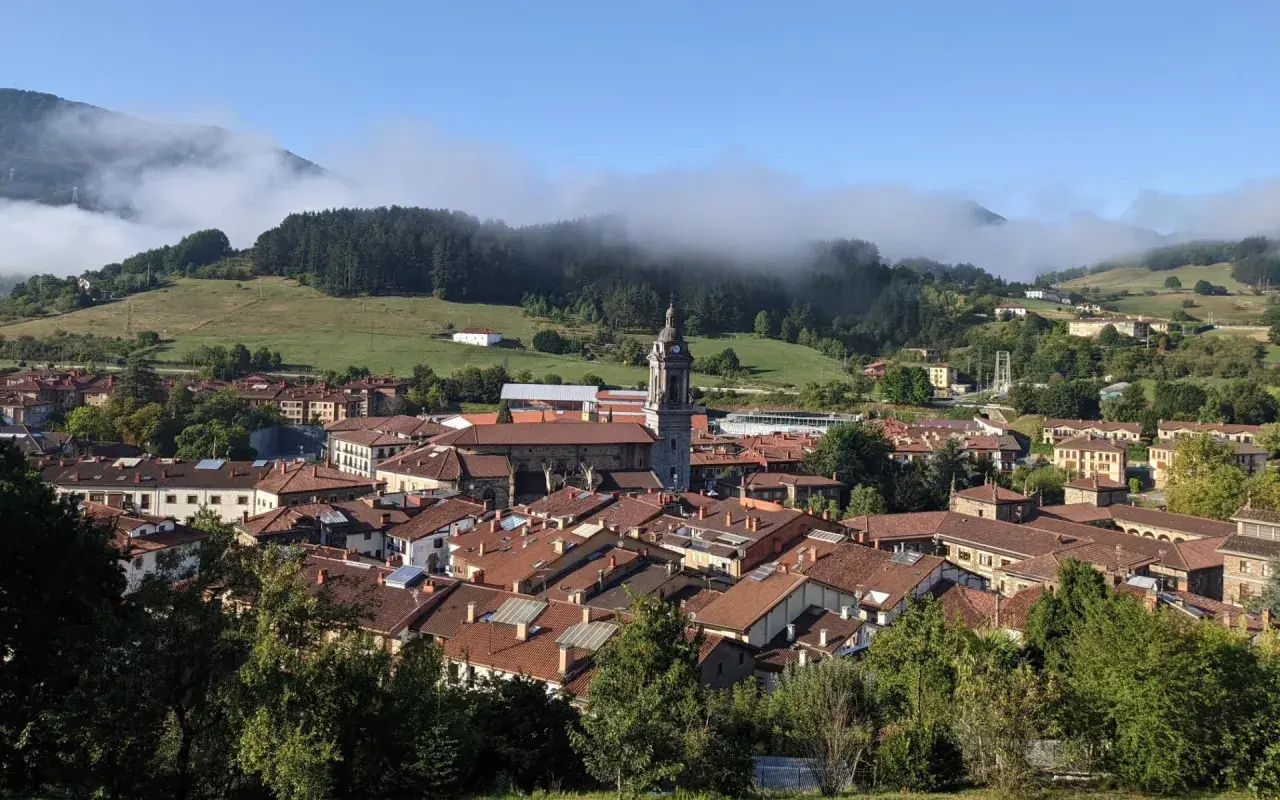 Vista panorámica de Oñati, con su iglesia y tejados rojizos, rodeada de verdes colinas y niebla. Ideal para planificar cómo llegar.