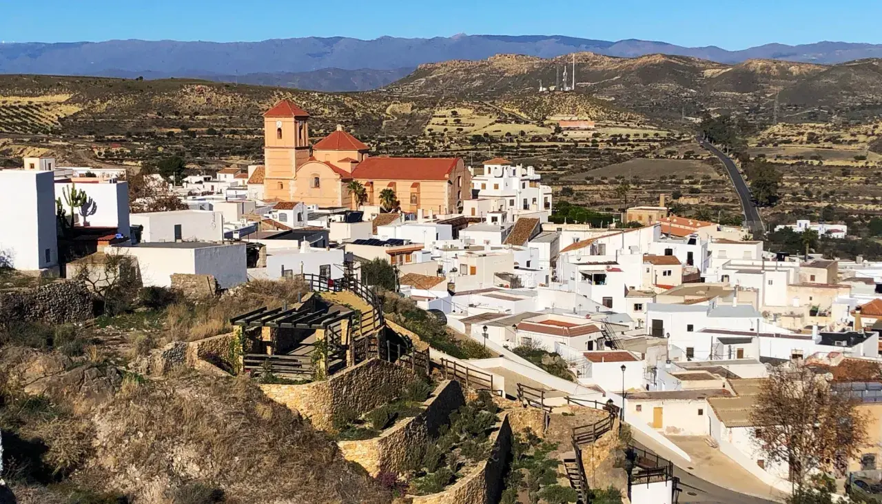 Vista panorámica de Lucainena de las Torres, un pueblo blanco andaluz con una iglesia de campanario naranja. El camino para llegar serpentea entre colinas áridas.