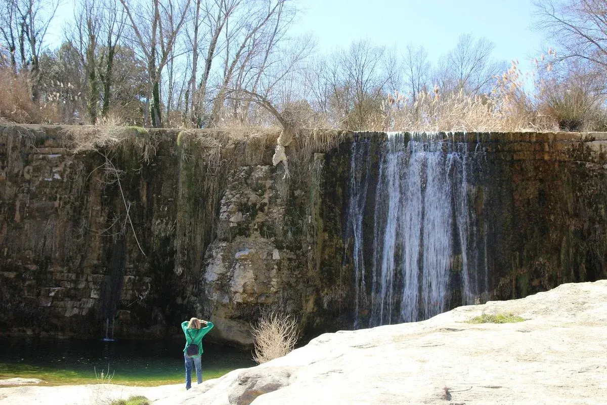Una persona admira una cascada en un paisaje rocoso. El agua cae sobre la pared de roca, creando un velo. El sol brilla, y la vegetación seca rodea la escena. Pozan de Vero, un lugar de belleza natural.