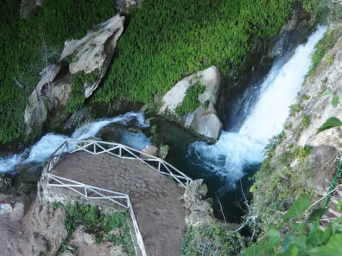 Sendero de piedra y barandilla blanca junto a la cascada de la cueva del agua Tiscar, rodeada de vegetación exuberante.