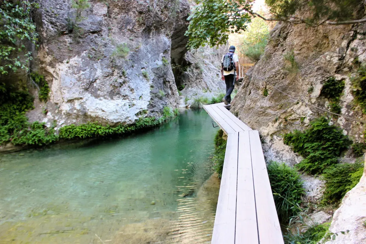 Senderista cruza un puente de madera sobre el agua cristalina en el Parrizal de Beceite, rodeado de rocas y vegetación exuberante.