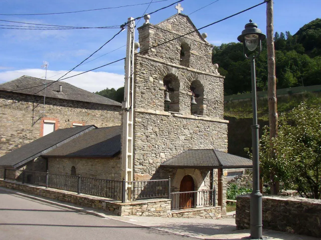 Iglesia de piedra en La Portela de Valcarce, con campanario de dos arcos y campanas. Un farol ilumina la entrada.