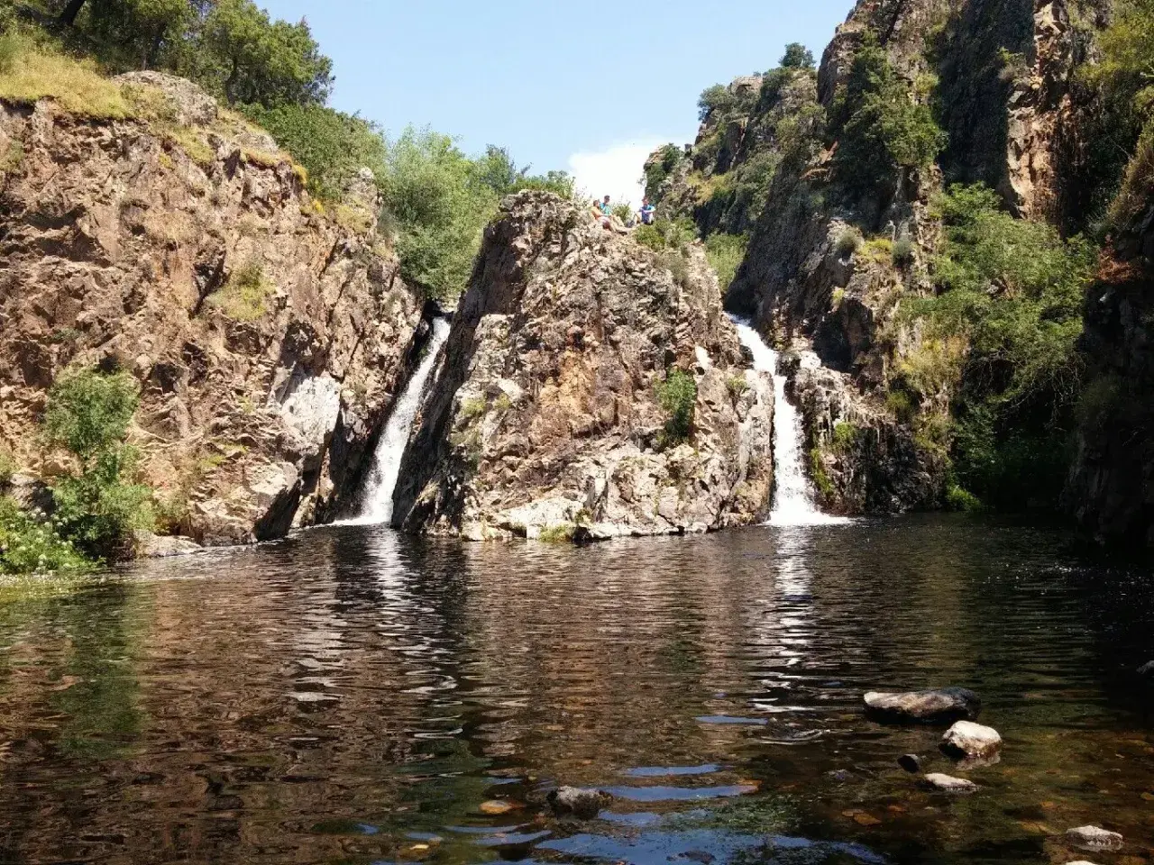 Dos cascadas caen por rocas escarpadas en la cascada del hervidero, formando un estanque oscuro. Personas observan desde lo alto.
