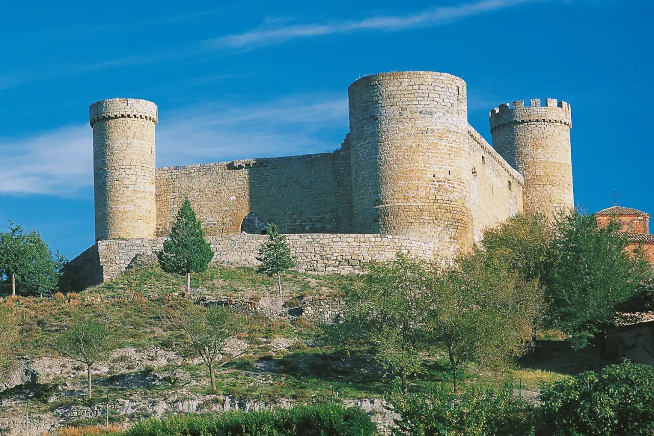 El castillo de Cornago se alza imponente sobre una colina, con sus torres circulares y murallas de piedra bajo un cielo azul intenso.
