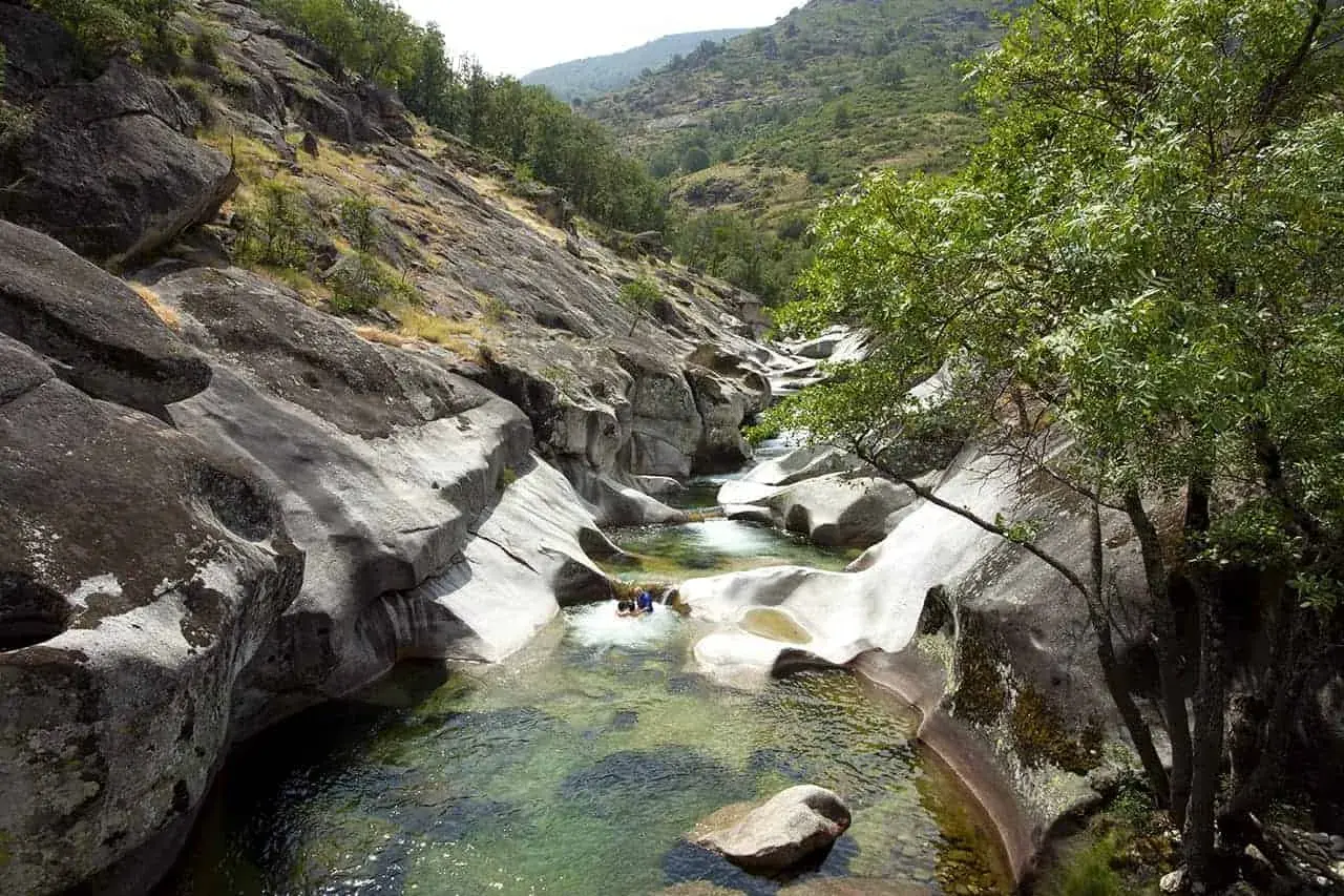 Un río cristalino serpentea entre rocas pulidas en la Garganta de los Infiernos. Dos personas disfrutan del agua fresca en este paisaje natural.