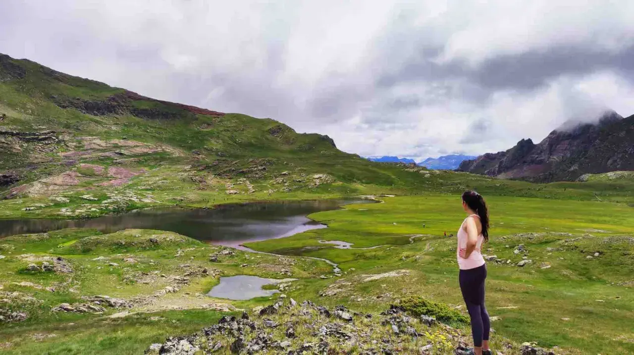 Mujer contempla los ibones de Anayet, lagos de montaña rodeados de verdes prados y picos rocosos bajo un cielo nublado.