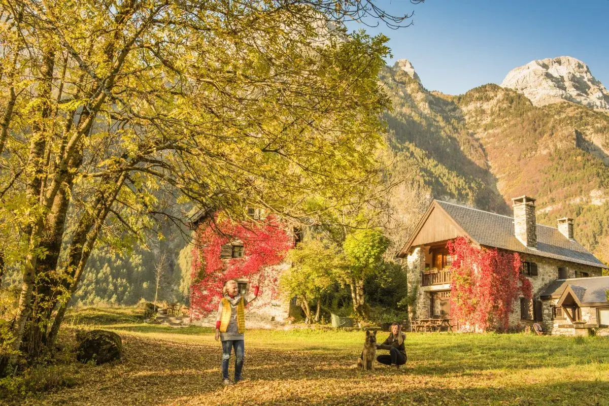 Dos mujeres y un perro disfrutan del otoño en el valle de Pineta, ideal para rutas. Las casas cubiertas de hiedra roja y las montañas crean un paisaje idílico.