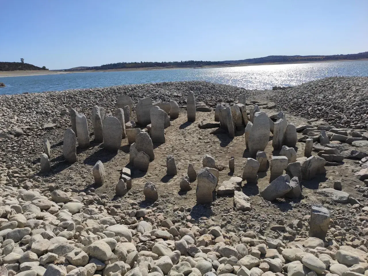 El dolmen de Guadalperal emerge de las aguas bajas, un círculo de menhires ancestrales sobre un lecho de piedras y arena, con un lago sereno al fondo.