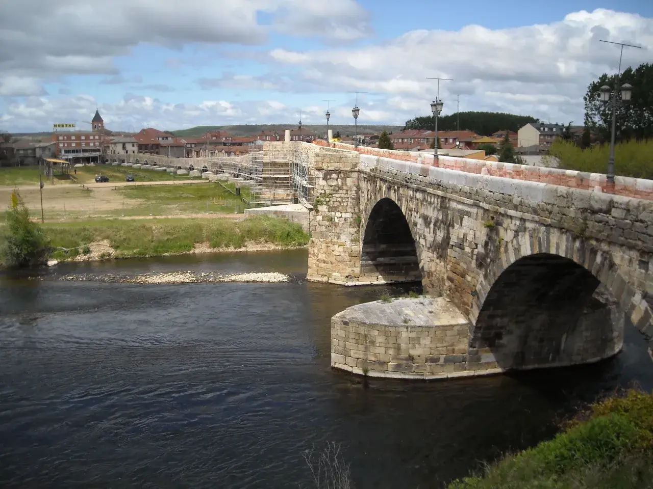 Puente romano en Hospital de Órbigo, con arcos de piedra y andamios. Al fondo, el pueblo y un hostal.