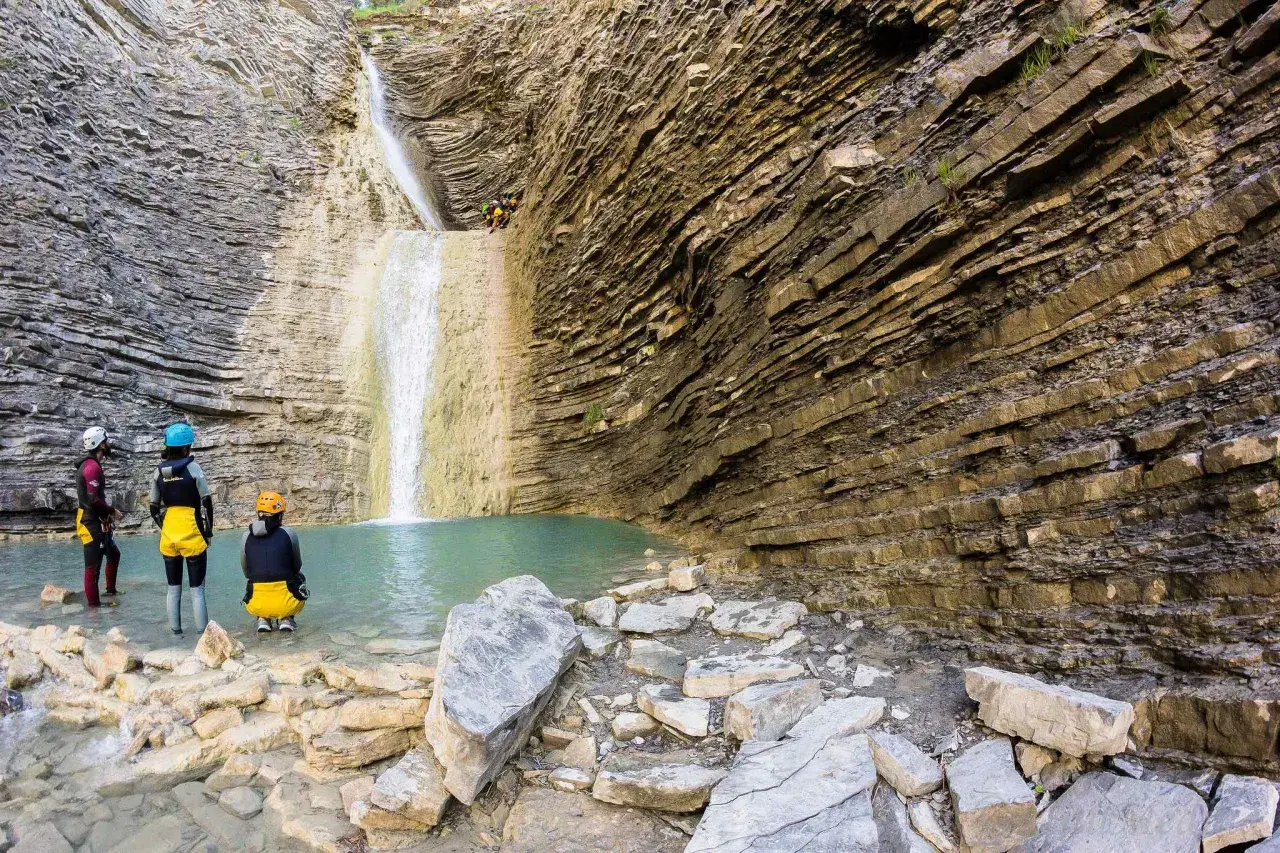 Un grupo de barranquistas se prepara para descender una cascada de oros bajo, rodeados de imponentes paredes rocosas estratificadas y una poza de agua turquesa.