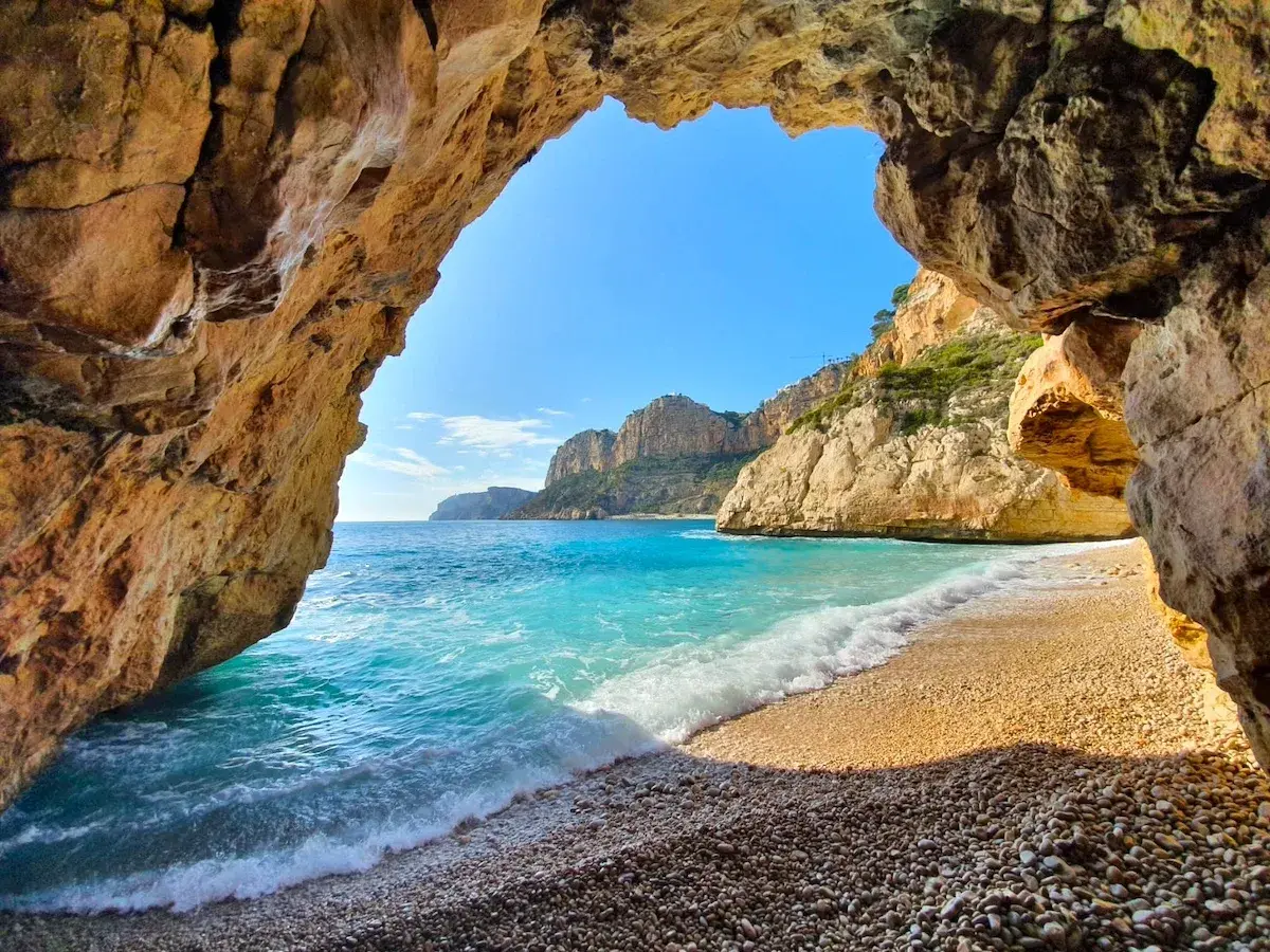 Vista desde una cueva en Cala Moraig, con aguas turquesas y acantilados dorados bajo un cielo azul.