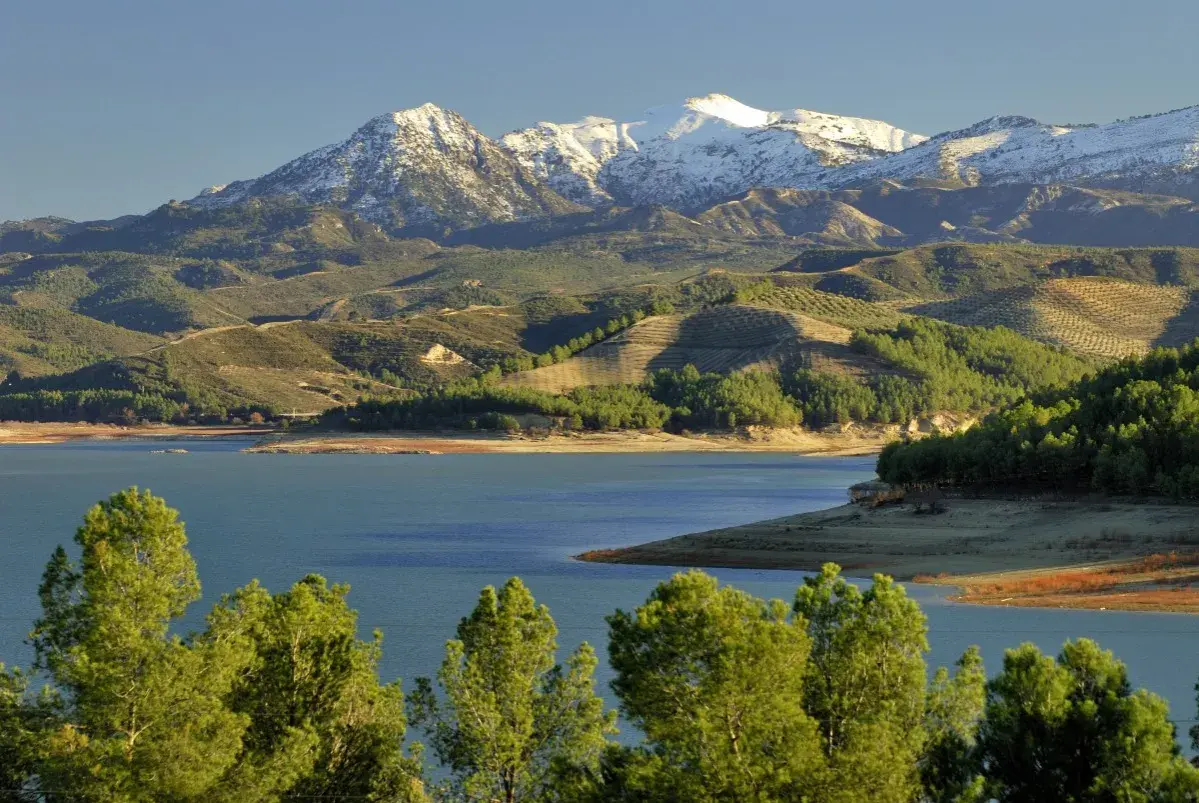 El pantano de los Bermejales, con montañas nevadas al fondo y colinas verdes salpicadas de olivos.