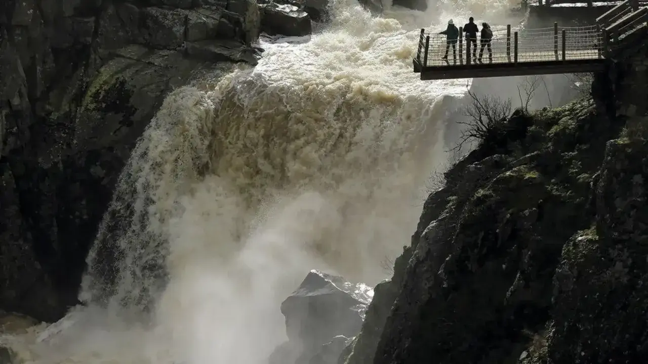 Tres personas observan la furia del pozo de los humos desde una pasarela. El agua turbia cae con fuerza, creando una densa niebla.