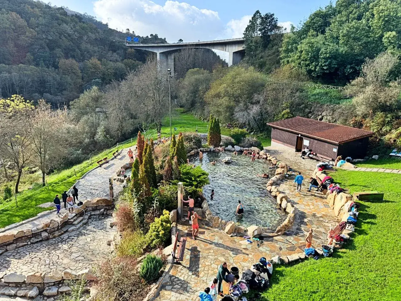 Gente disfrutando de las **termas en Galicia**, un oasis de relajación rodeado de naturaleza, con un puente moderno al fondo.