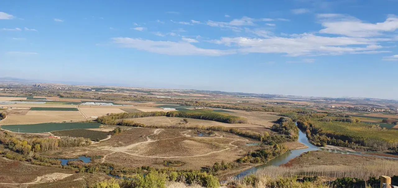 Paisaje rural con un río serpenteante cerca de Villafranca del Bierzo. Campos cultivados y árboles de otoño bajo un cielo azul con nubes.
