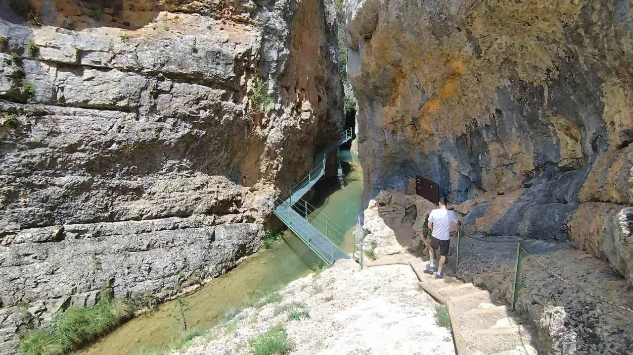 Un hombre camina por las pasarelas de Calomarde, un sendero metálico que serpentea sobre un río entre imponentes paredes rocosas.