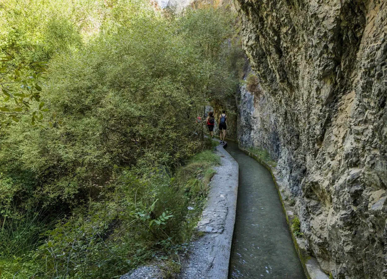 Senderistas caminan por la ruta de los bolos, un sendero estrecho junto a un canal de agua y un acantilado rocoso.