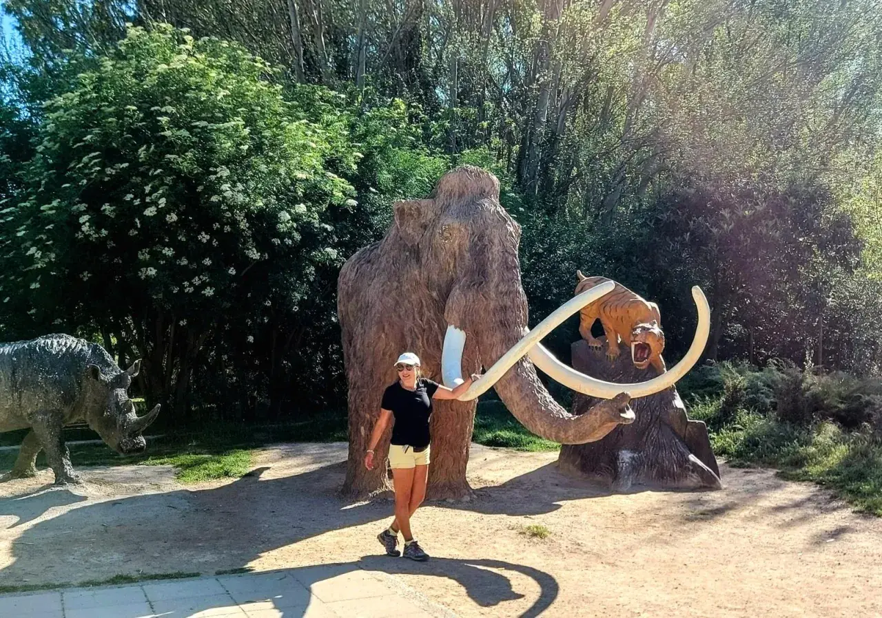 Una mujer sonríe junto a un mamut y un tigre dientes de sable, parte de la ruta del mamut. Un rinoceronte se ve a la izquierda.