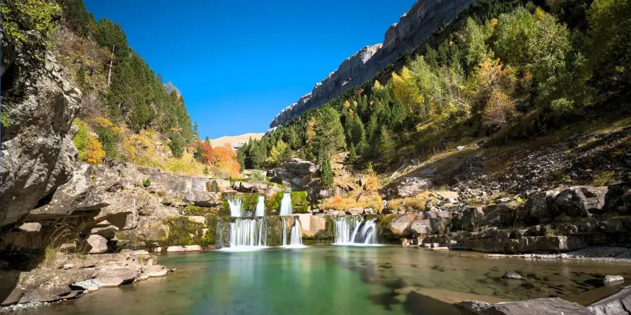 Cascada en un valle montañoso con árboles otoñales, ideal para las mejores rutas senderismo España.