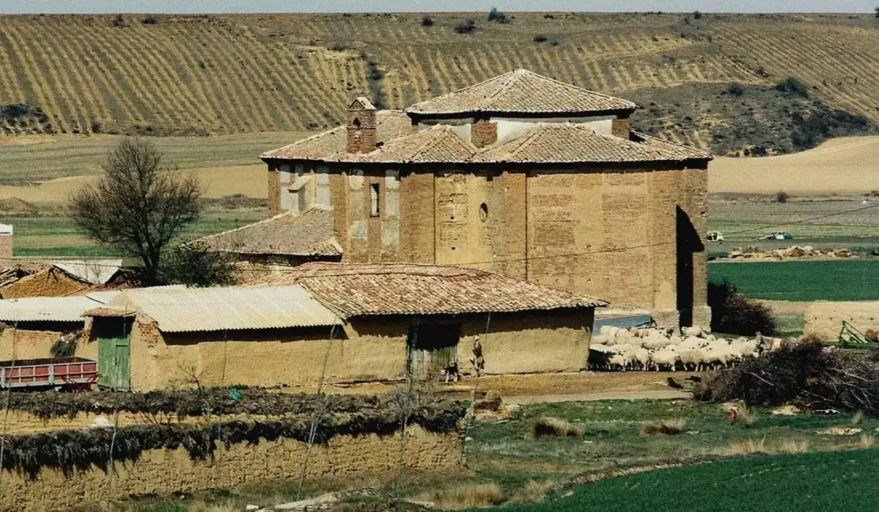 Edificio de ladrillo con tejado a dos aguas, rebaño de ovejas pastando cerca. Paisaje rural con campos de cultivo en terrazas, típico de calzadilla de la cueza.