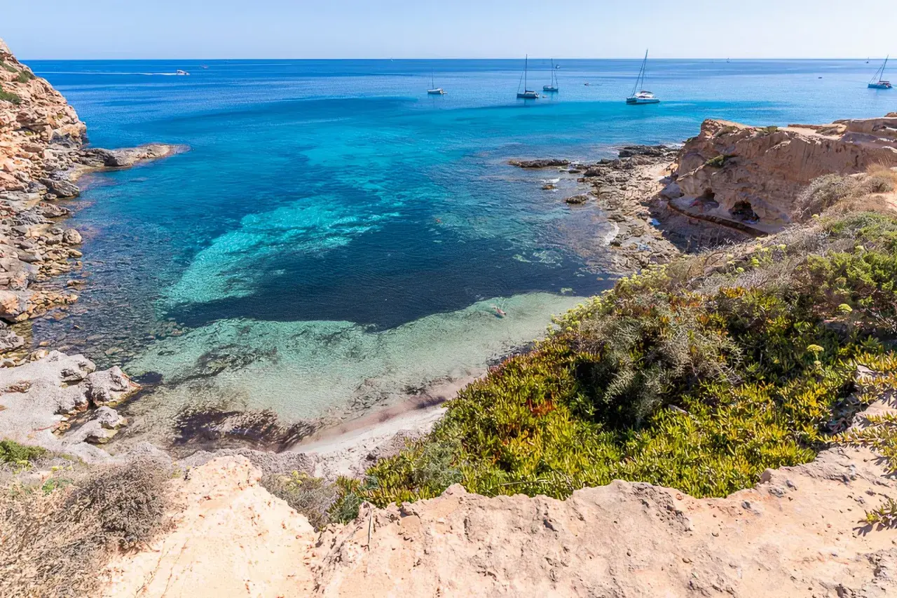 Aguas turquesas y arena blanca en una cala rocosa, un paraíso entre las mejores playas de Formentera. Yates navegan en el horizonte.