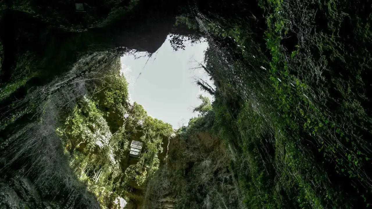Vista desde abajo del pozo de los aines, con paredes cubiertas de vegetación exuberante y cascadas de agua cayendo hacia el cielo abierto.