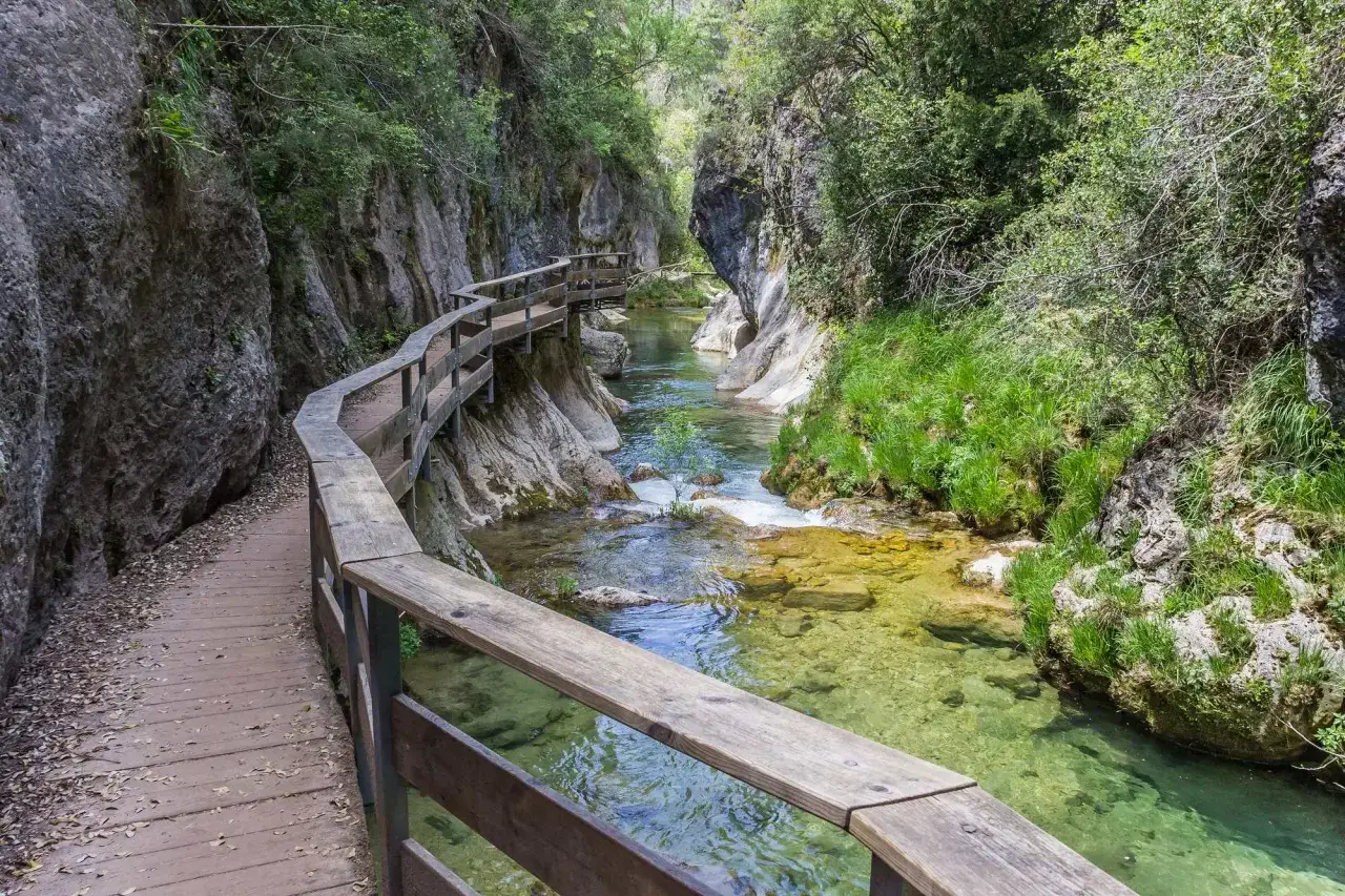 Pasarela de madera junto a r&iacute;o cristalino en ca&ntilde;&oacute;n, ideal para las mejores rutas senderismo Espa&ntilde;a.