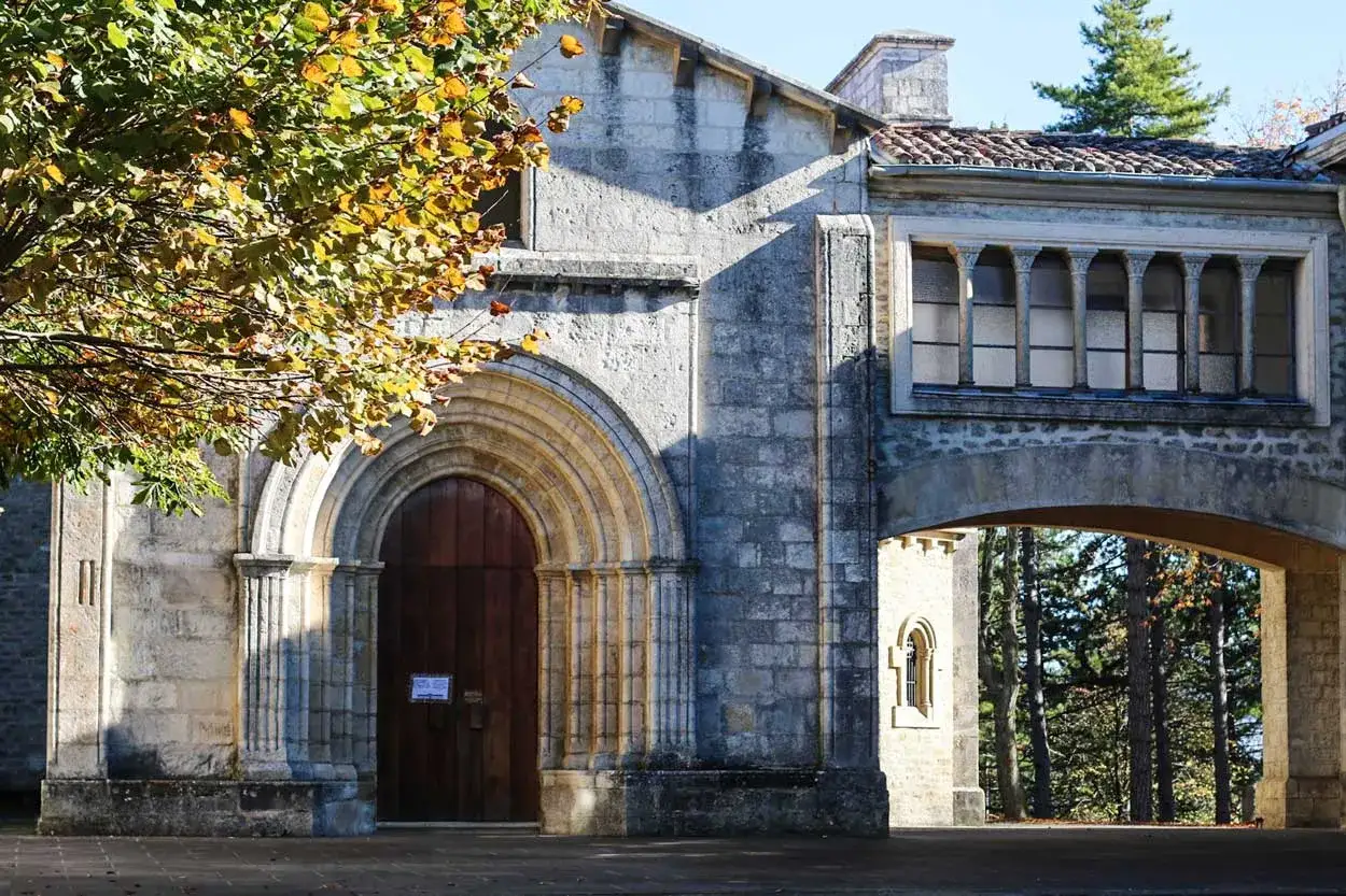 Fachada del santuario de Estíbaliz con un arco de piedra y puerta de madera. Un árbol con hojas otoñales ilumina la escena.